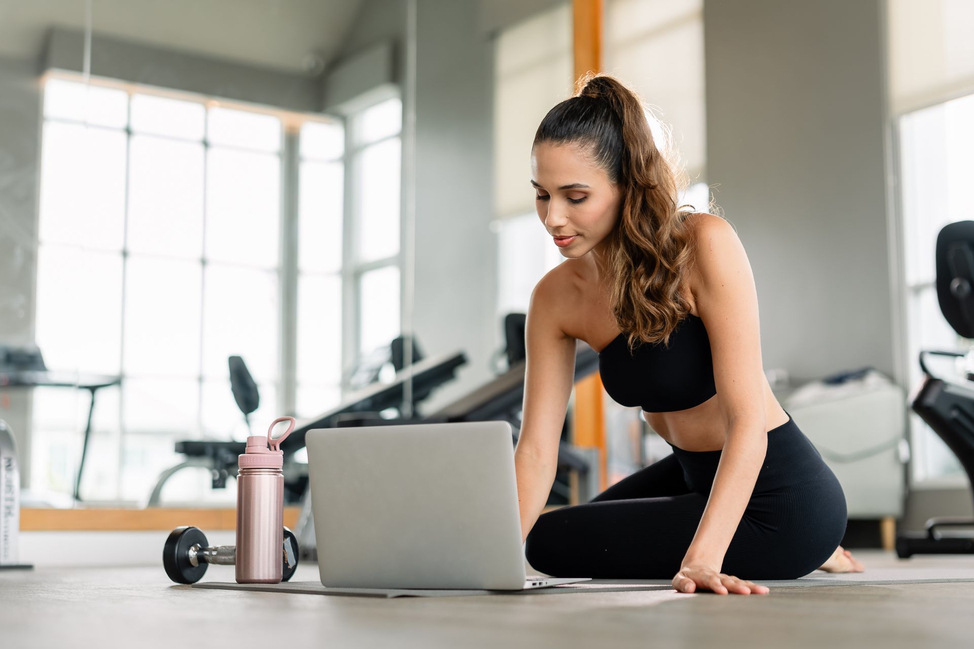 Woman in gym clothes using laptop on the floor in a gym setting.