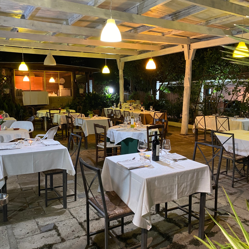 Patio esterno del ristorante con tovaglie bianche, tavoli e sedie sotto un pergolato con luci sospese.