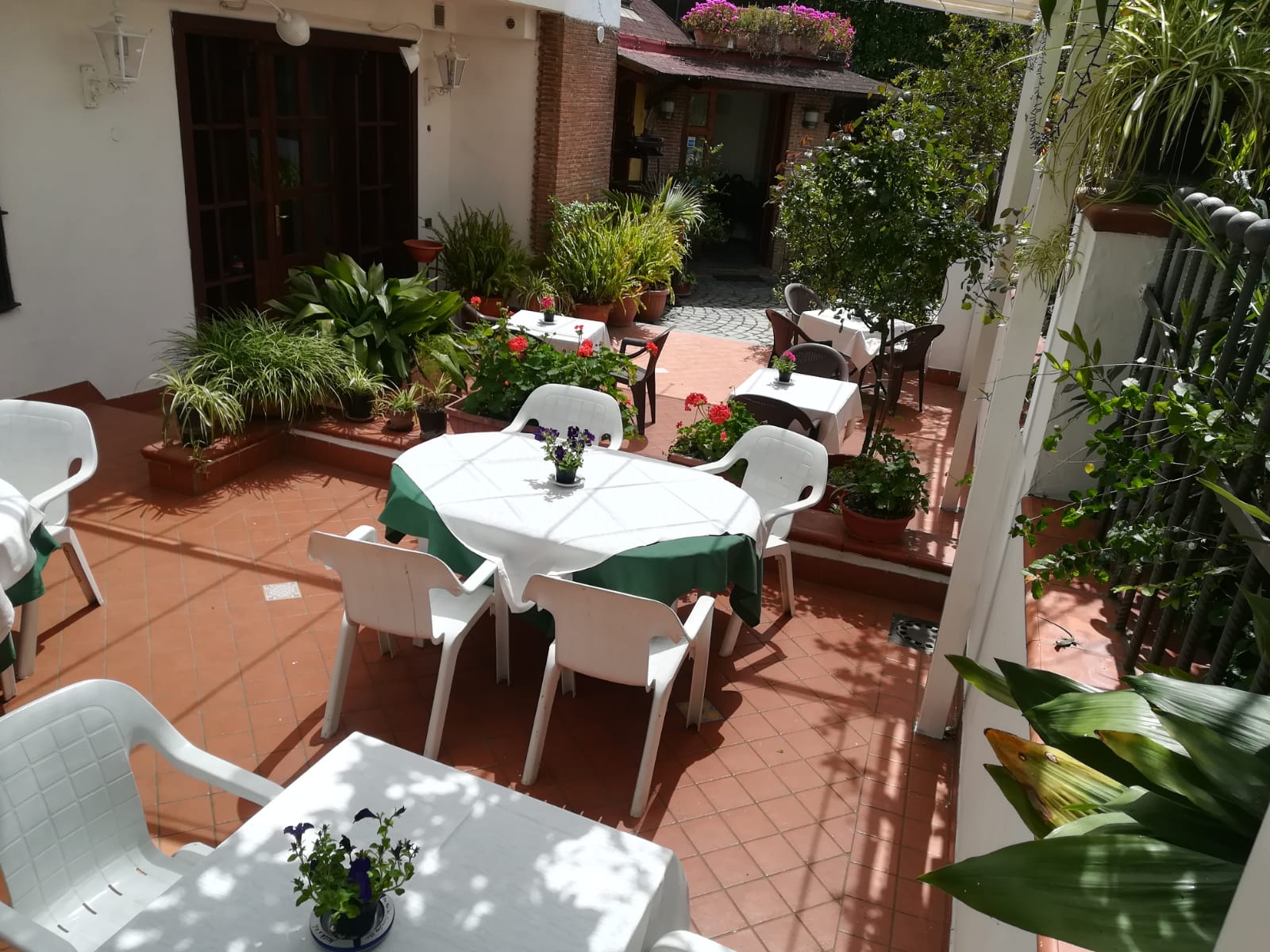 Patio esterno con tavoli bianchi, sedie e tovaglie verdi, circondato da piante e da un edificio.