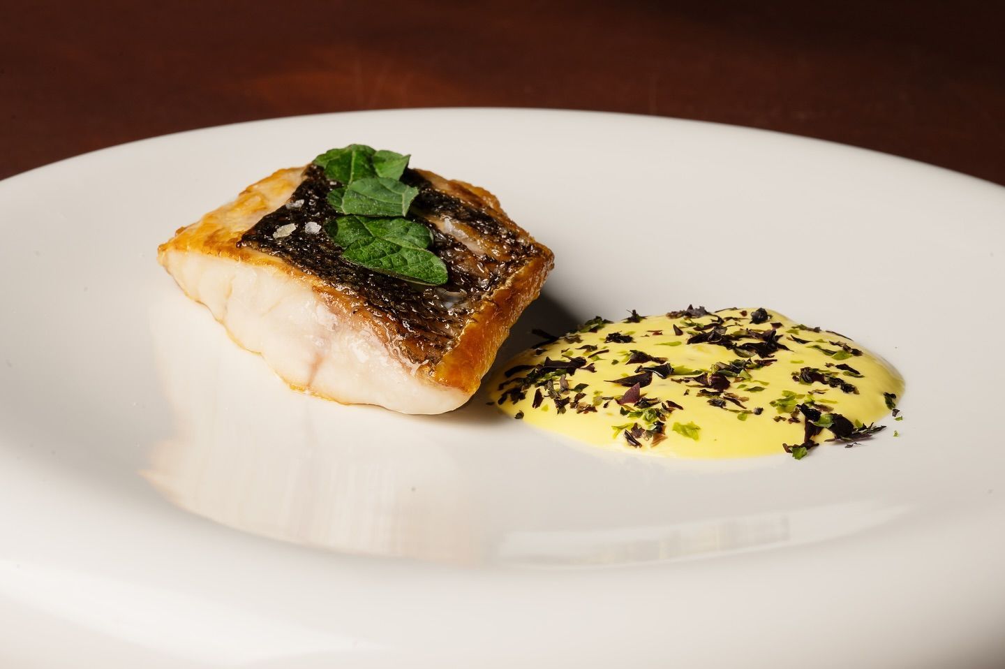 Filetto di pesce scottato in padella con salsa gialla, guarnito con erbe verdi su un piatto bianco.