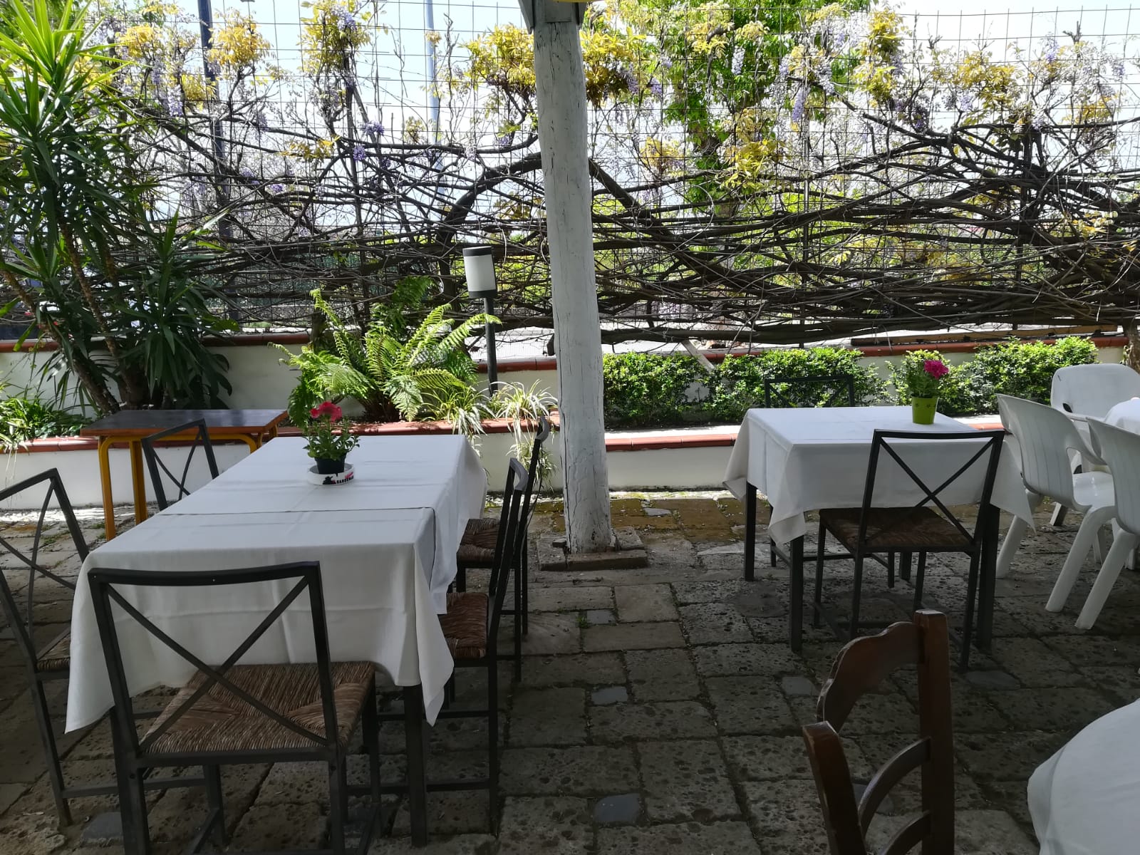 Patio esterno del ristorante con tavoli, sedie e fiori, racchiuso da una recinzione decorativa con vegetazione.
