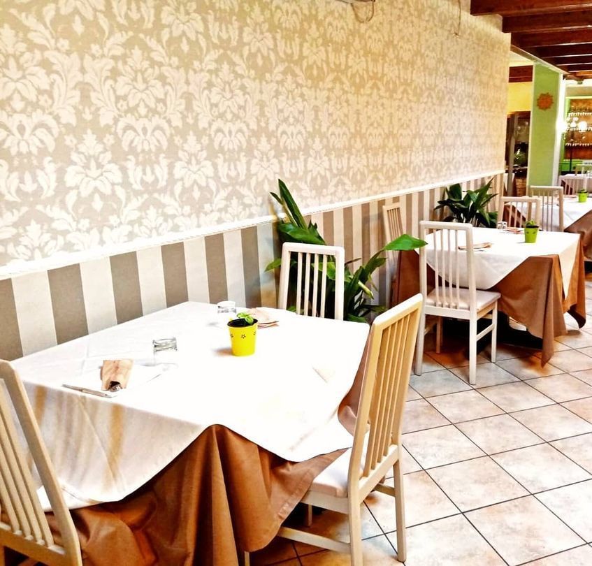 Interno di un ristorante con tavoli apparecchiati per la cena, tovaglie marroni, sedie color crema e carta da parati decorativa.