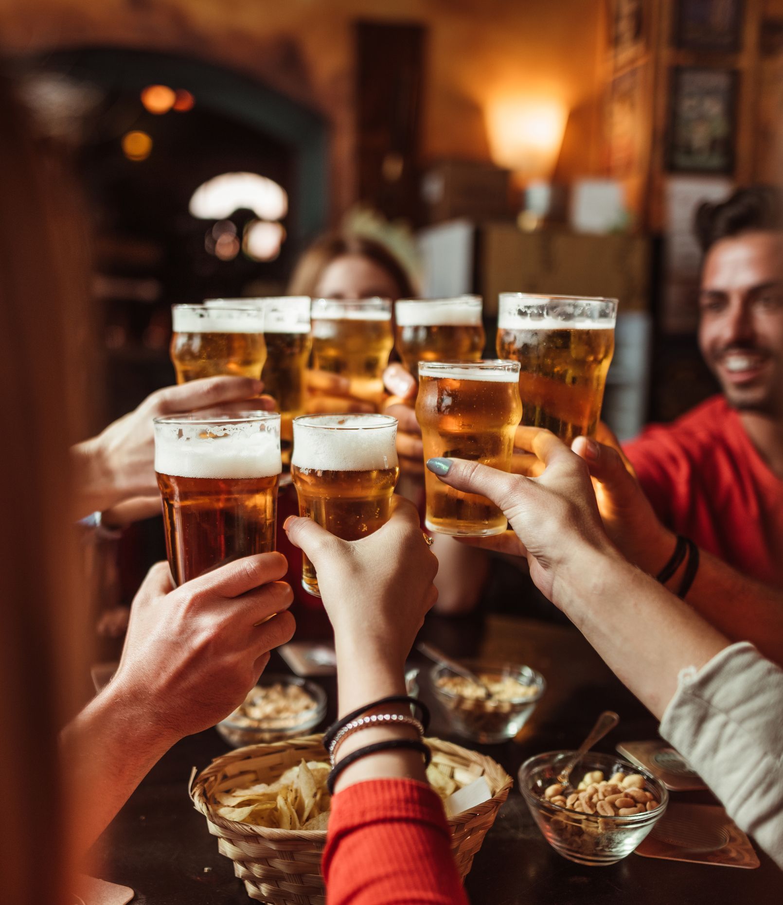 Persone che brindano con pinte di birra in un pub. Espressioni allegre, luci calde. Stuzzichini sul tavolo.