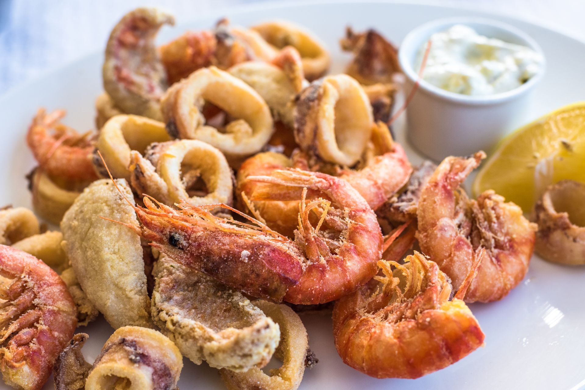 Piatto di pesce fritto, tra cui gamberi e calamari, con contorno di salsa bianca e uno spicchio di limone.