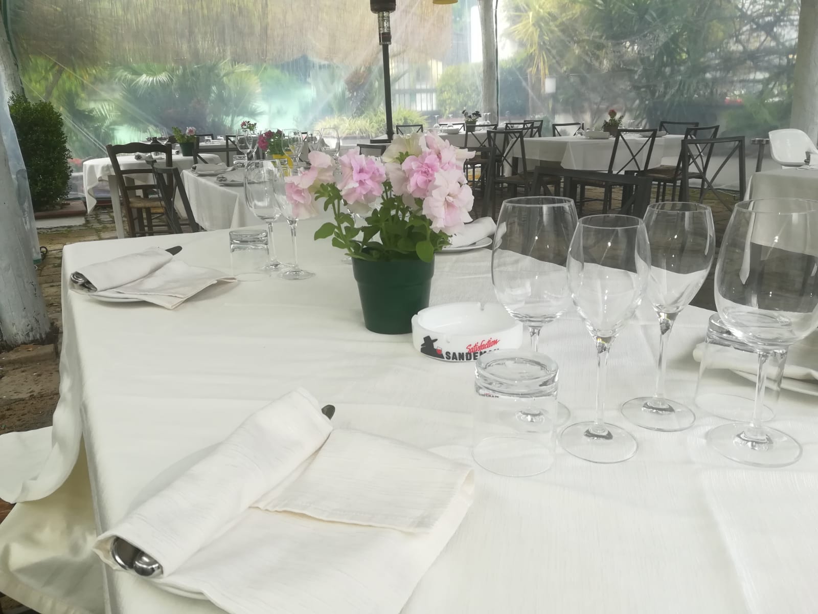 Tavolo da pranzo per esterni con tovaglia bianca, bicchieri, tovaglioli e fiori rosa.