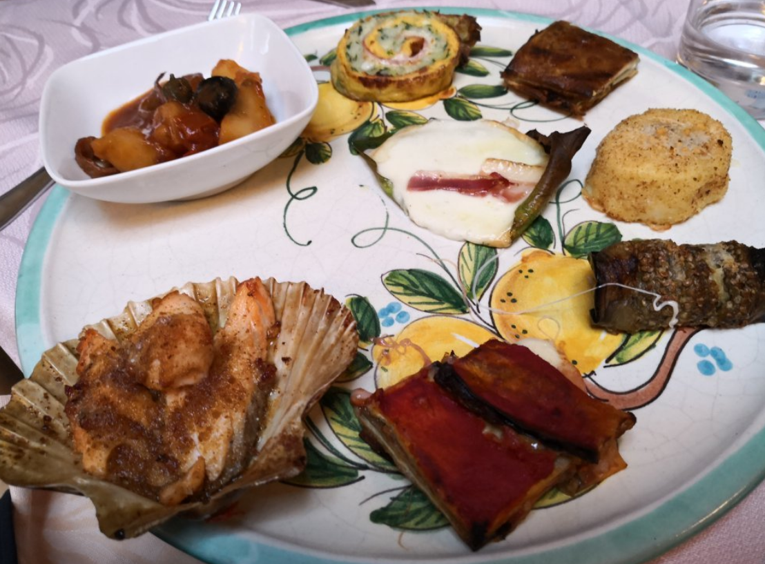 Un piatto di antipasti: capesante, verdure e involtini, presentati su un piatto decorato con fiori.