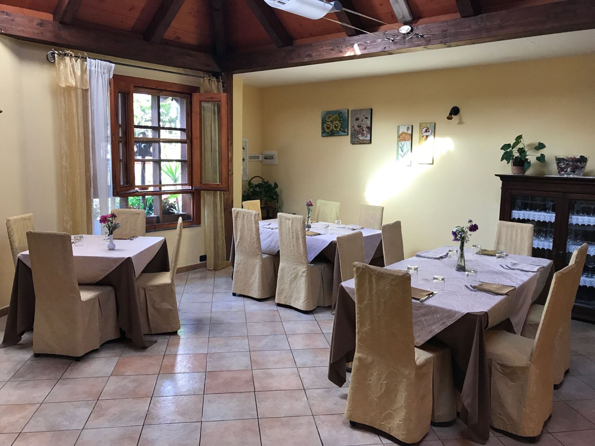 Sala da pranzo del ristorante con tavoli apparecchiati per i pasti, sedie e tovaglie color crema, finestra e mobiletto decorativo.