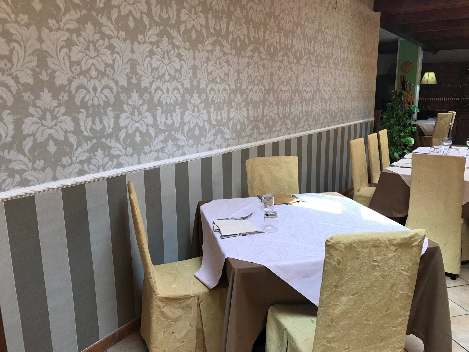 Interno del ristorante con tavoli apparecchiati per cenare; carta da parati a motivi e rivestimenti a righe.
