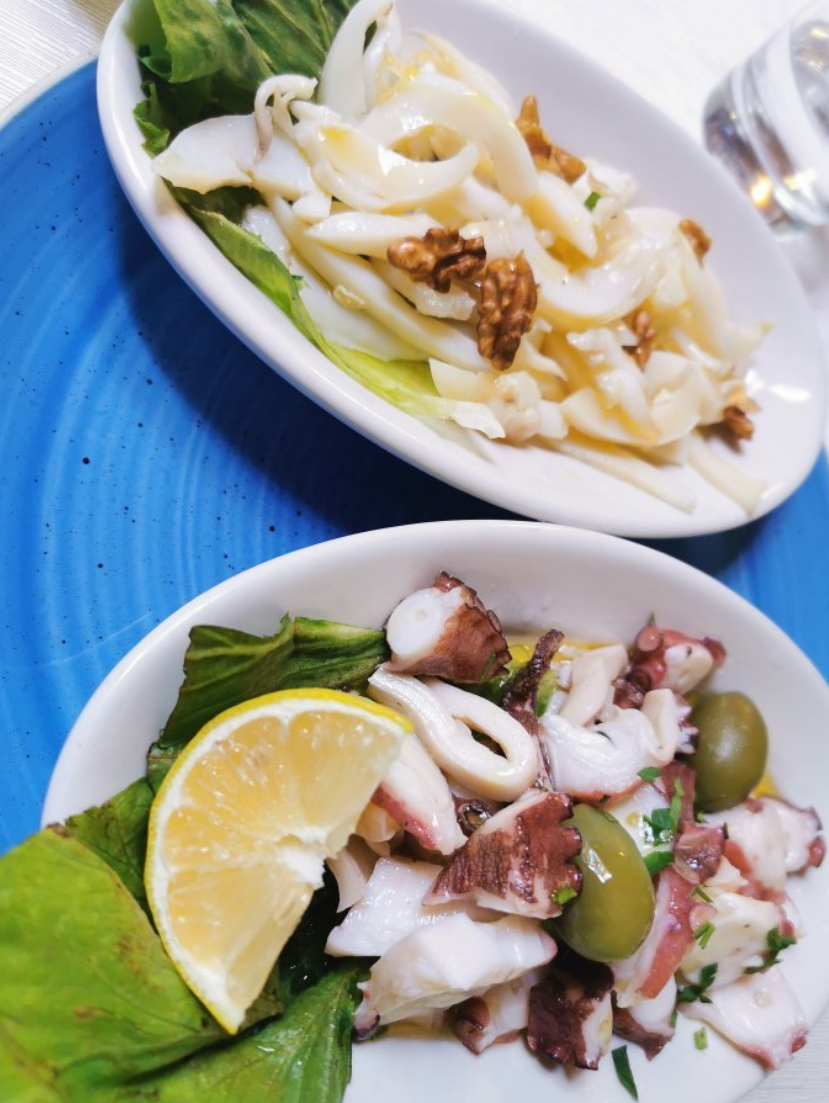 Due antipasti di mare: insalata di calamari e polpo su piatti bianchi, sfondo blu.