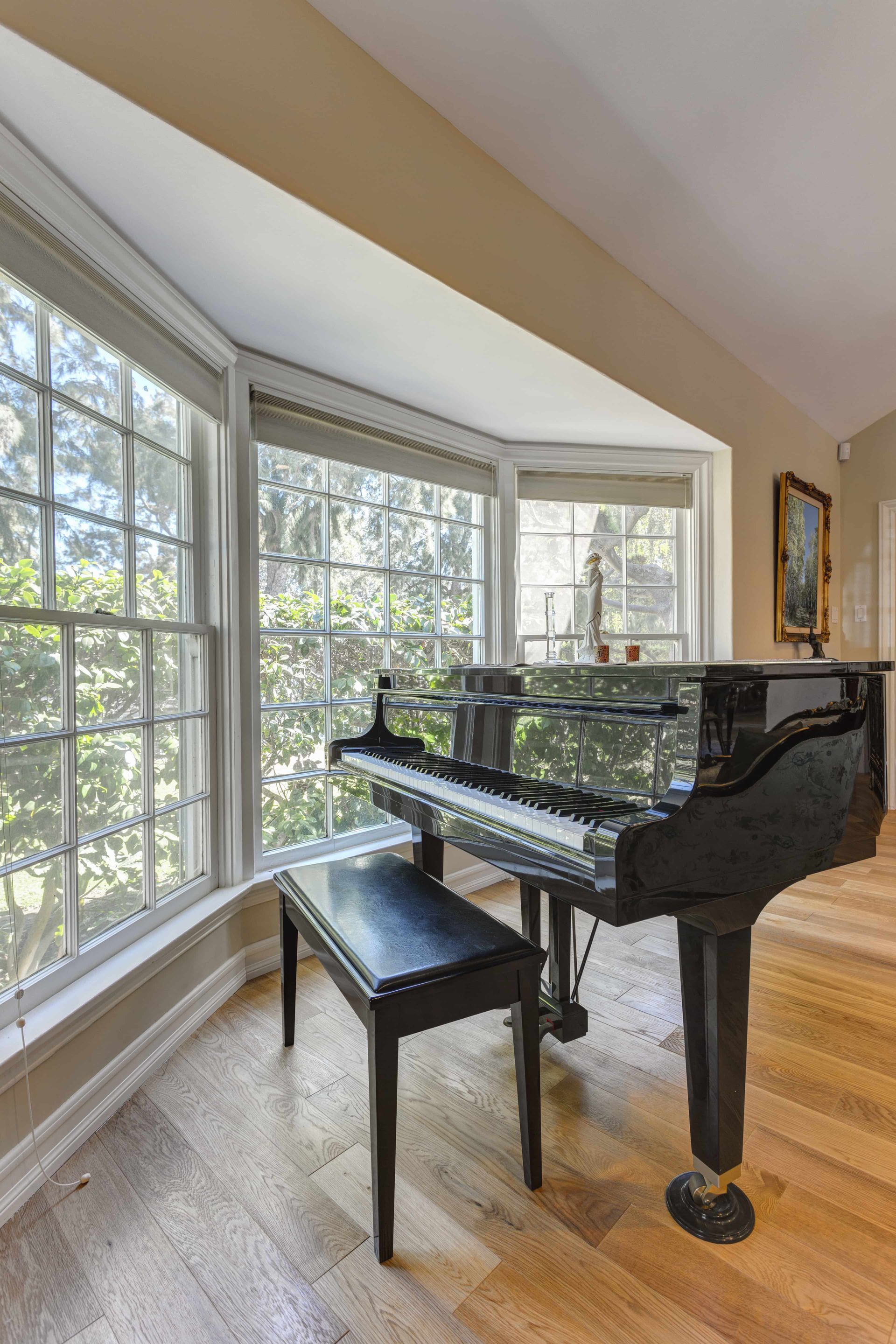 A black grand piano with a matching bench placed by a large bay window in a room with hardwood floors.