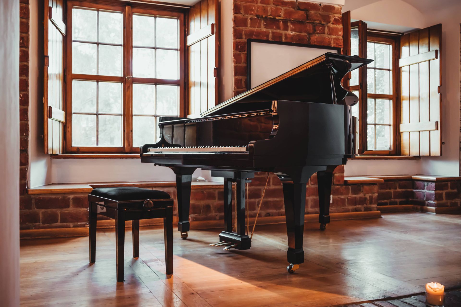 A black grand piano with a matching bench in a sunlit room with brick walls and large windows.