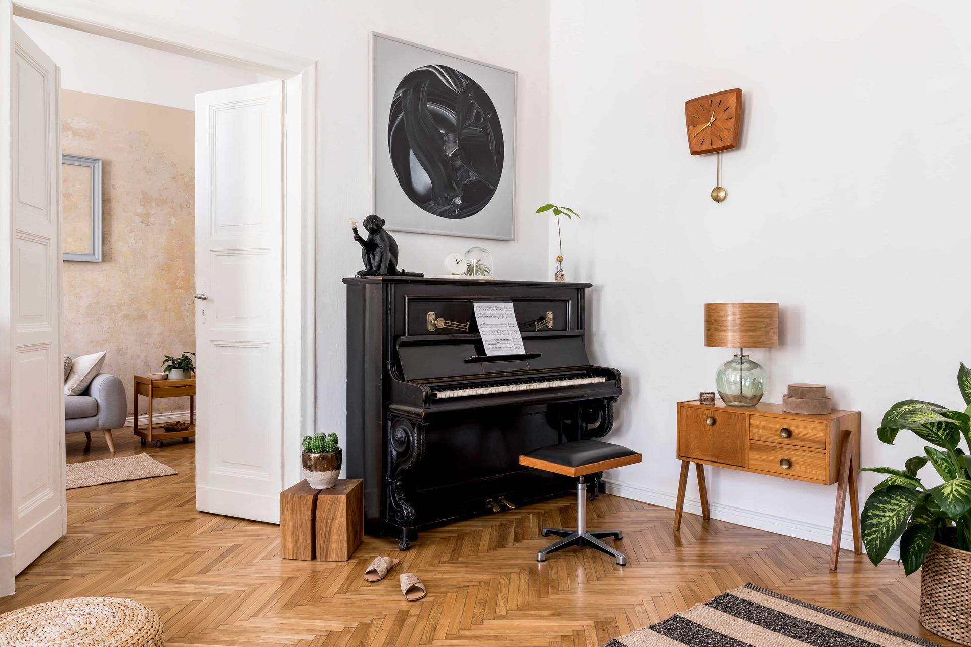A black upright piano with a stool sits in a brightly lit room with herringbone wood floors, wall art, and decor.