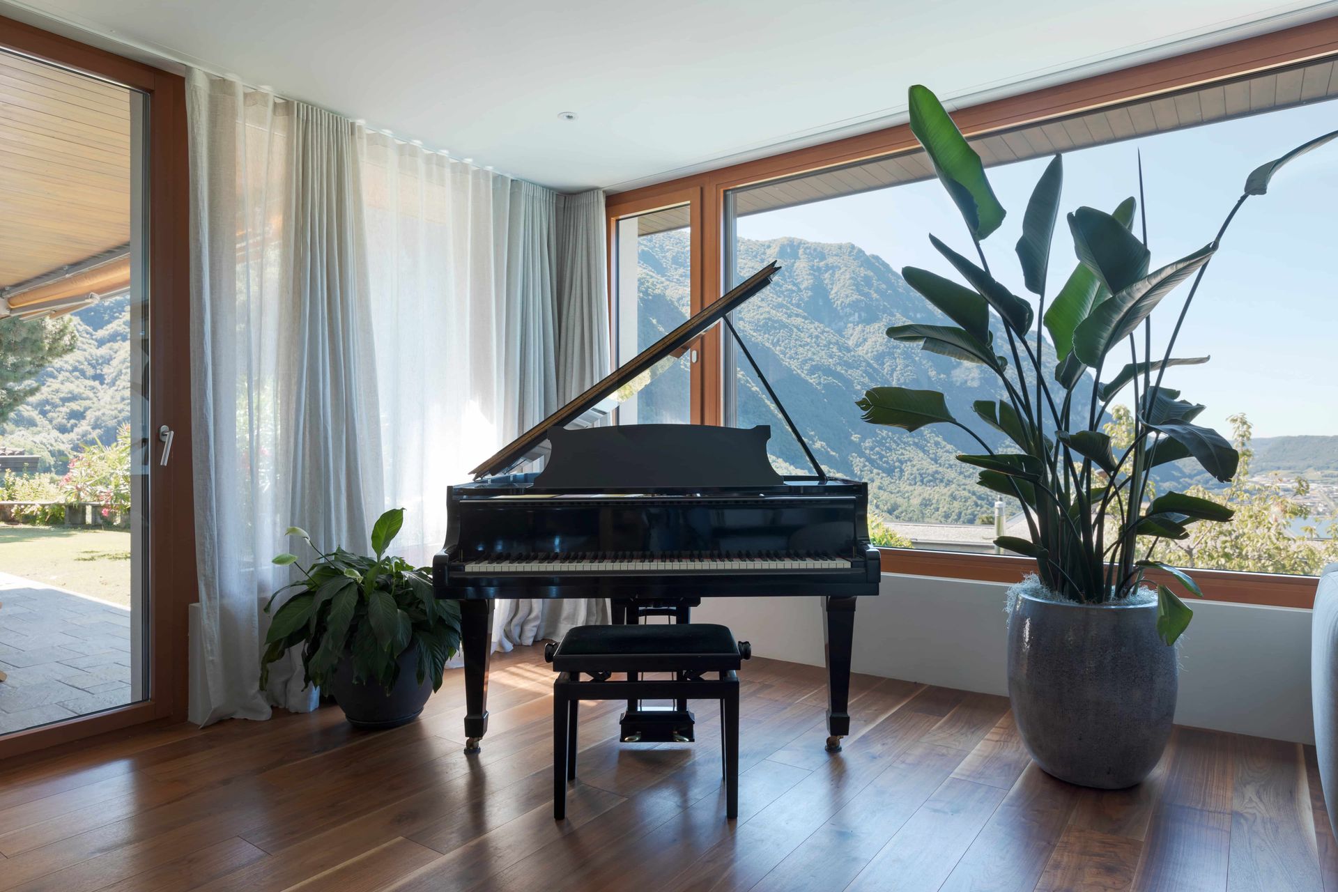 A grand piano sits on a wooden floor in a sunlit room with large windows, sheer curtains, and two large potted plants.