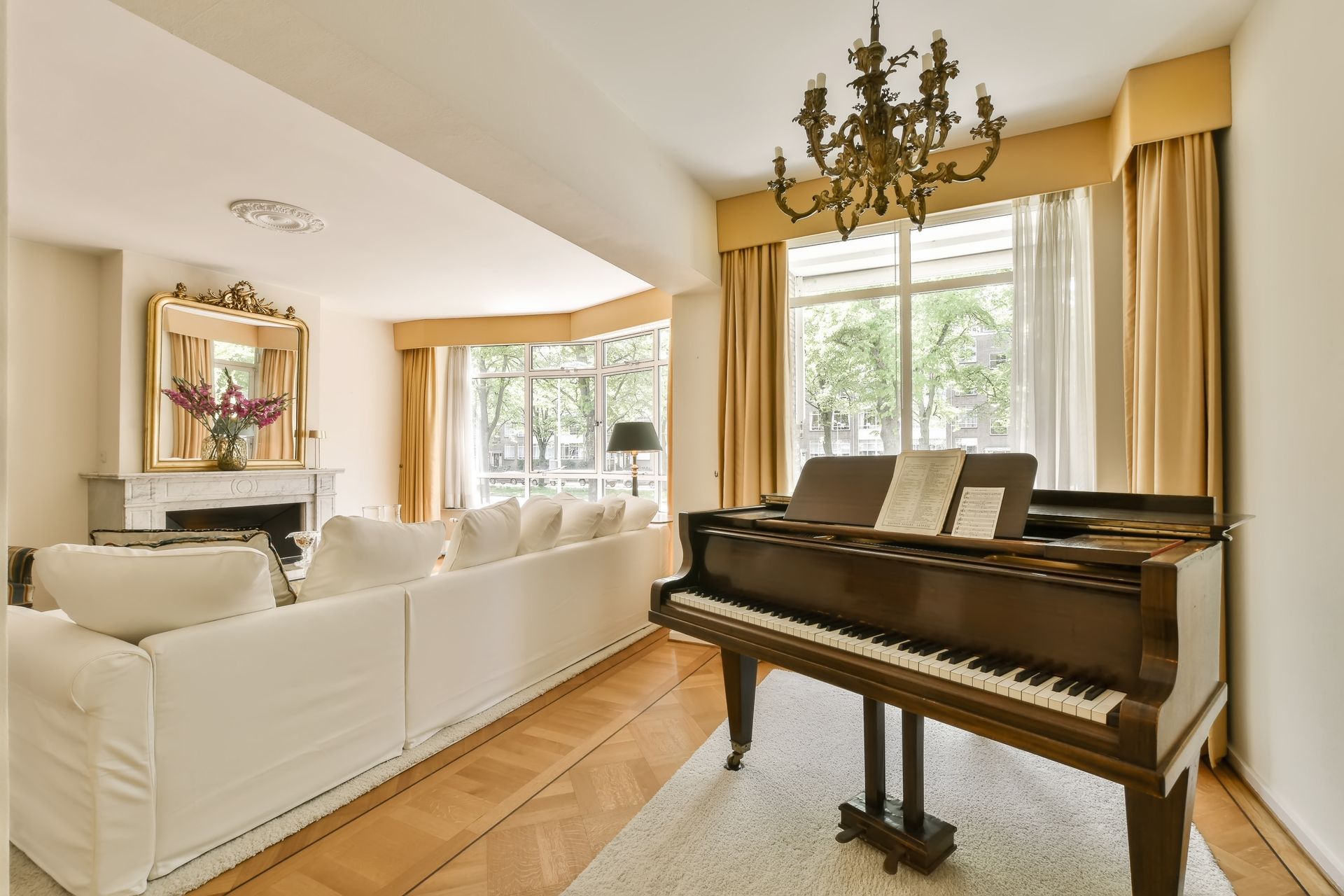 A sunlit living room featuring a grand piano, a white sofa, and a fireplace with a large mirror in a classic interior.