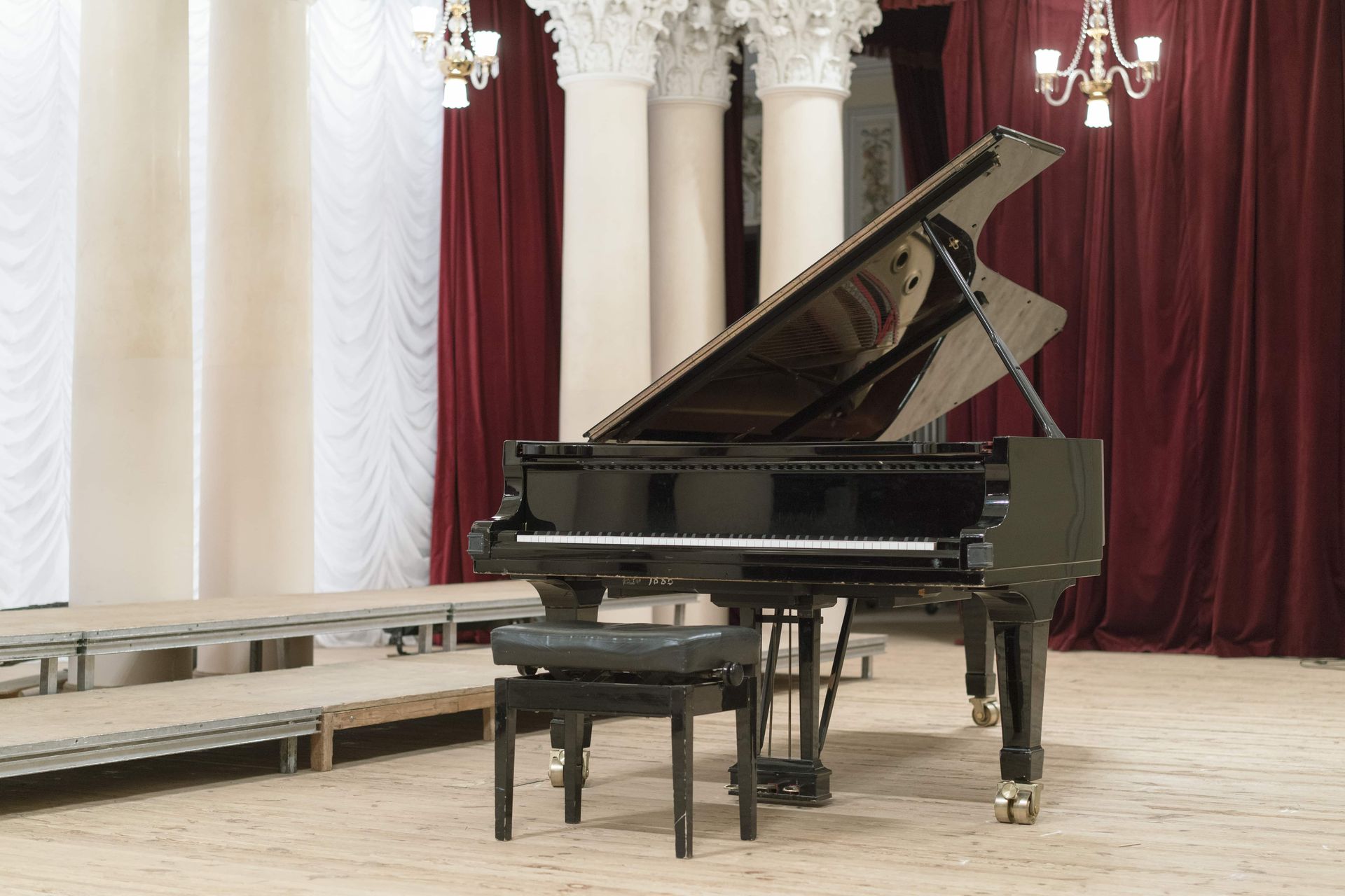 A black grand piano with a matching bench sits on a wooden stage in front of white columns and dark red curtains.
