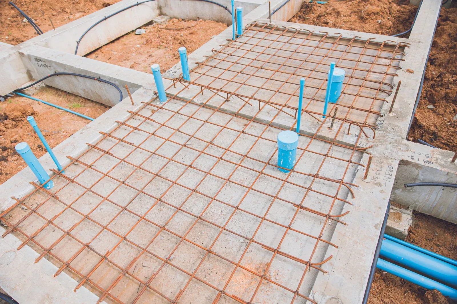 Foundation construction with rebar grid, blue pipes, and concrete formwork.