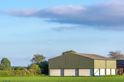 Green barn with a concrete base, set in a green field under a blue sky with clouds.