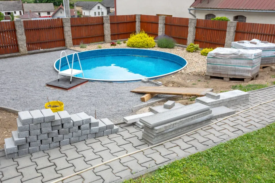 Backyard pool under construction with paving stones, gravel, and wooden fence.