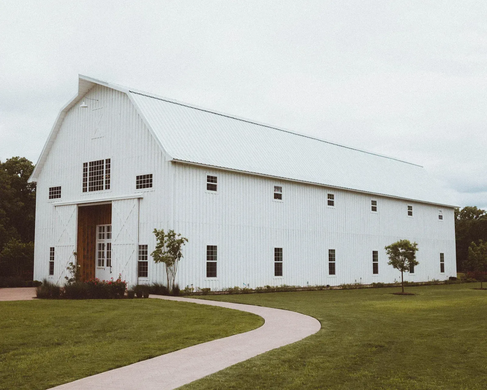 White barn with a curved path leading to the entrance, trees and green grass on a cloudy day.