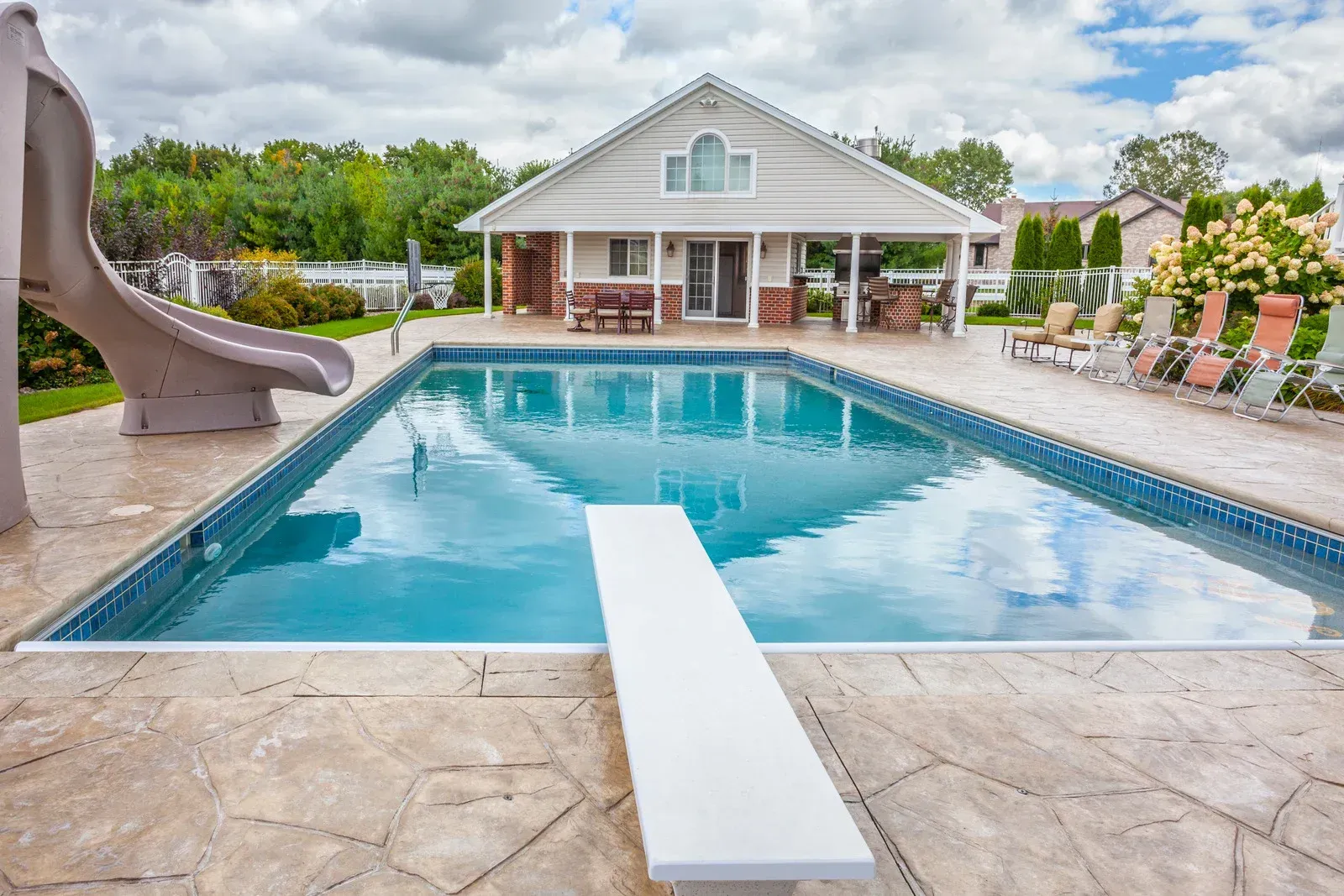Swimming pool with diving board and slide, a small house in the background, and lounge chairs along the side.