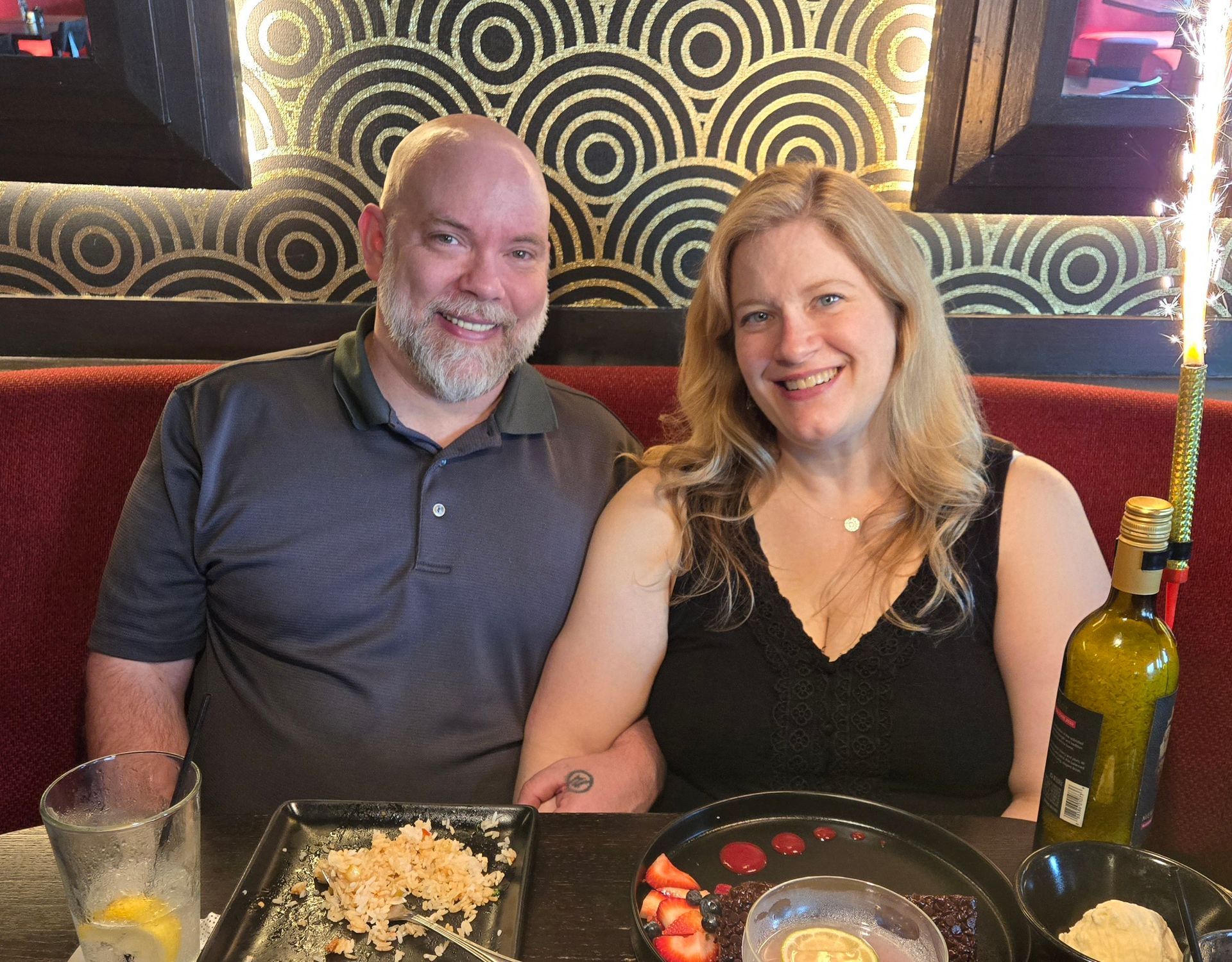 Couple at restaurant; smiles at camera. Dessert plate, wine bottle, fountain sparkler, gold and black decor.