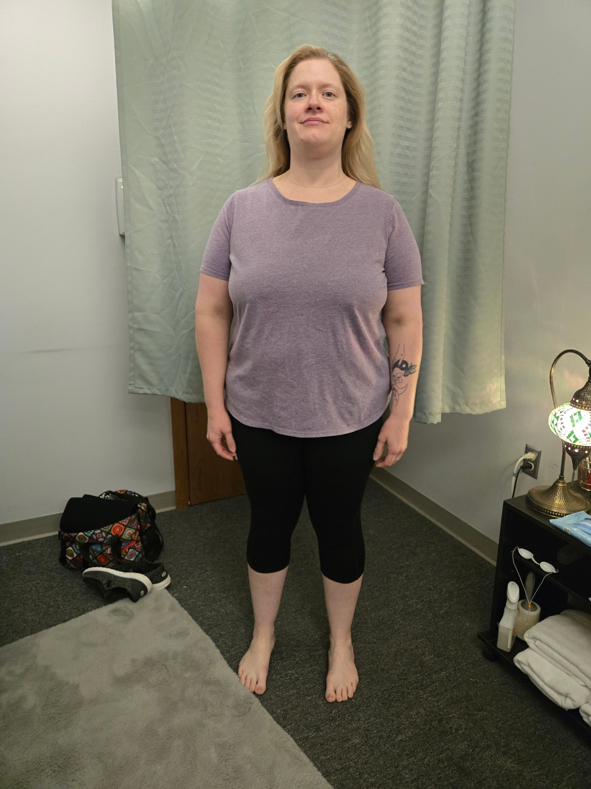 Woman standing indoors, wearing purple shirt and black capris, smiling.