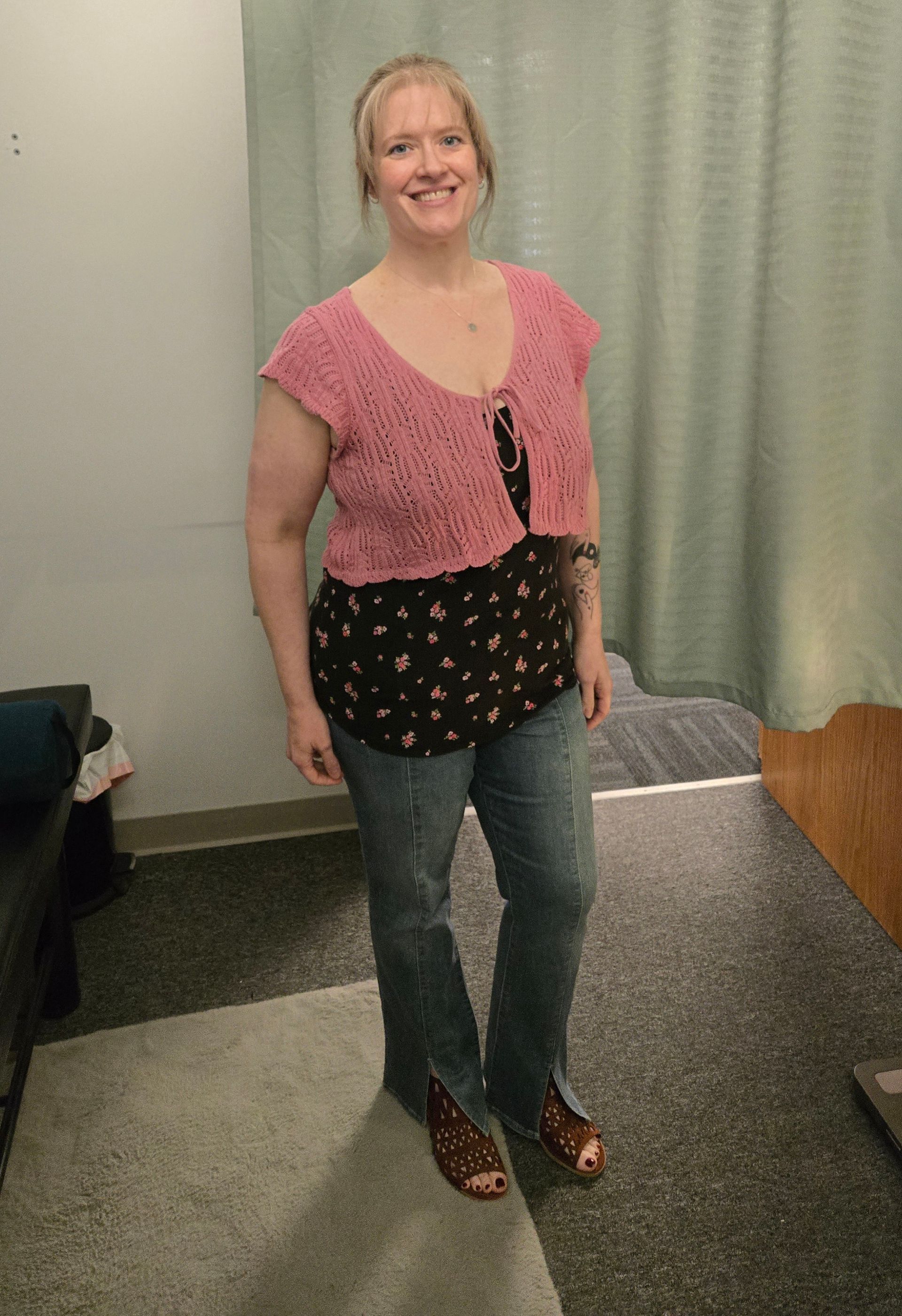 Woman wearing pink top, black floral shirt, and jeans, smiling in a room.