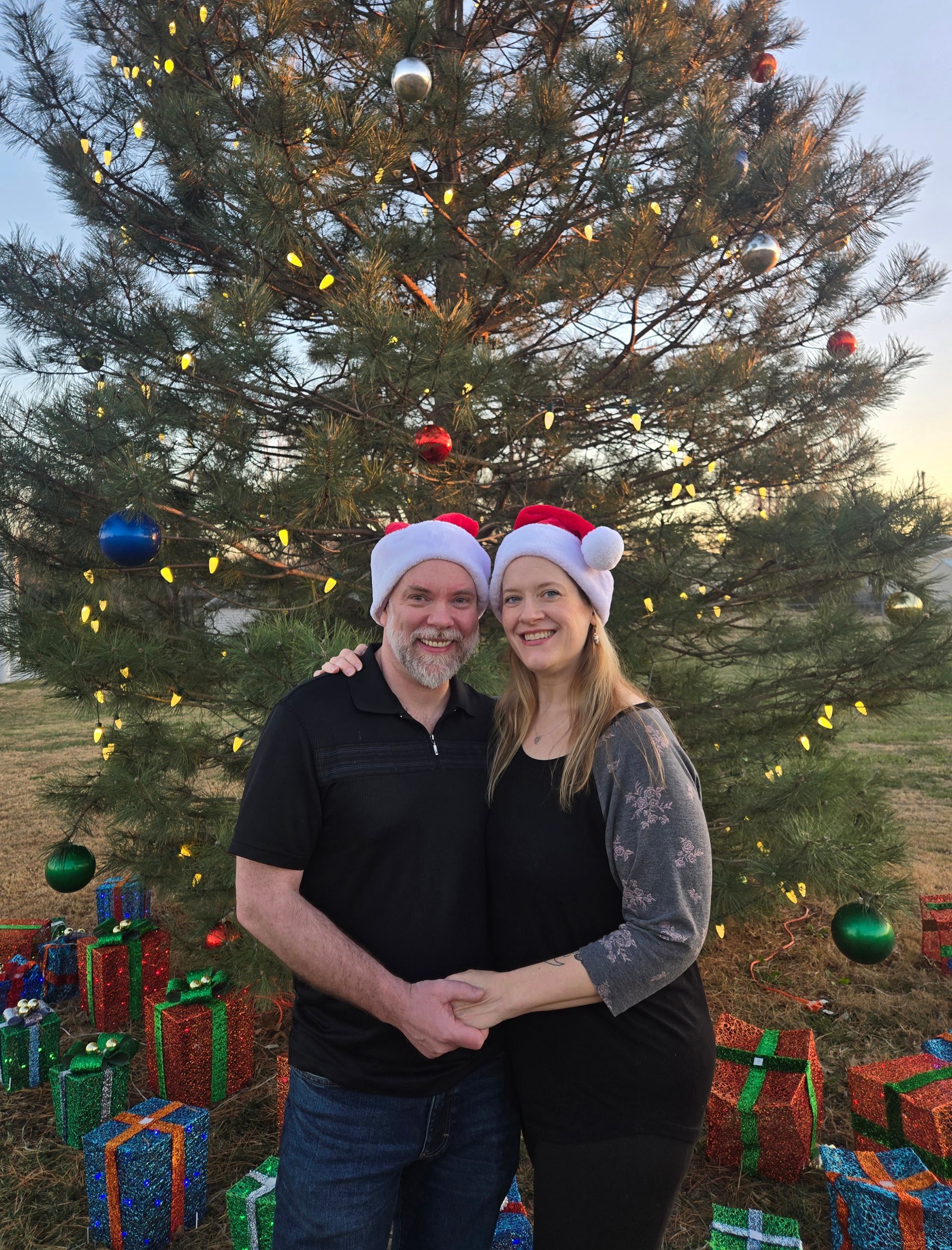 Couple holding hands in front of Christmas tree, wearing Santa hats, surrounded by wrapped presents.