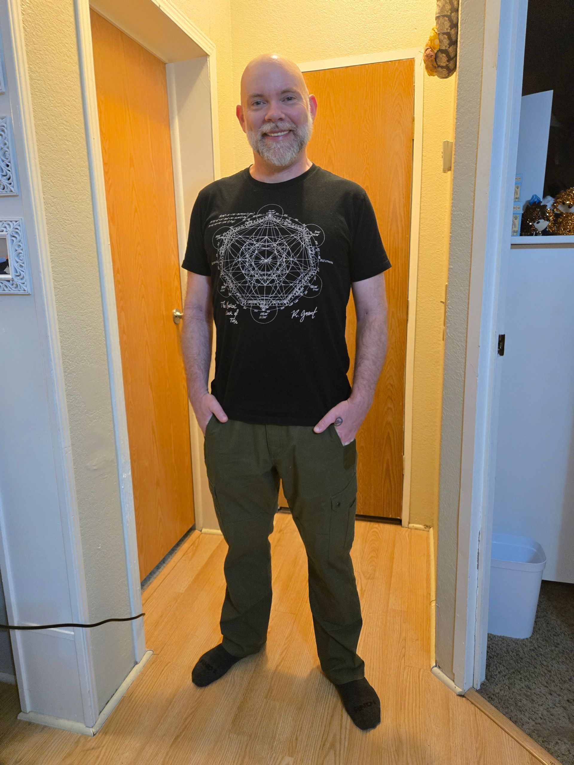 Man in black t-shirt and green pants stands in doorway, smiling. Hands in pockets.