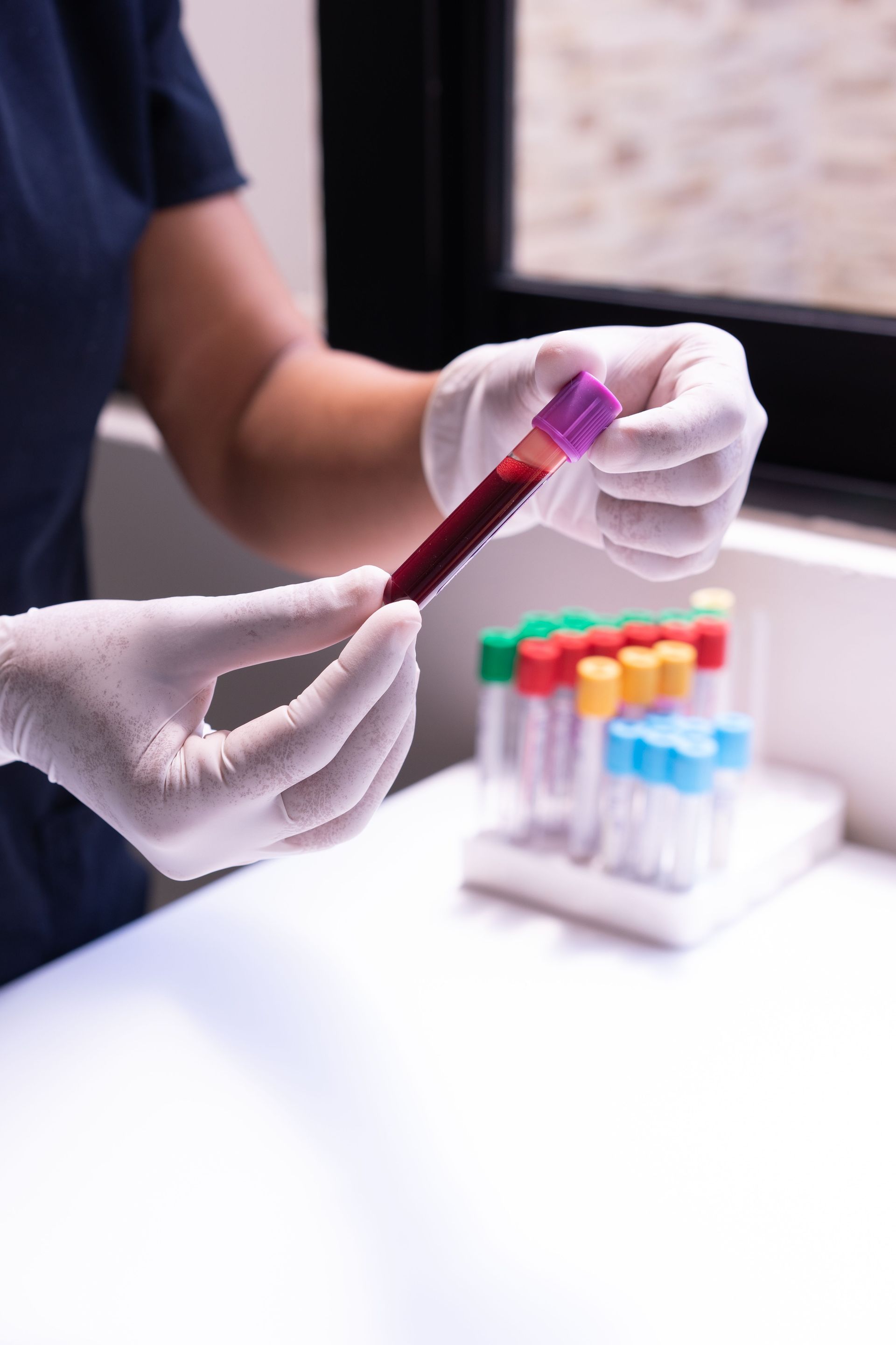 Gloved hands hold a blood sample tube beside a rack of colorful test tubes in a lab
