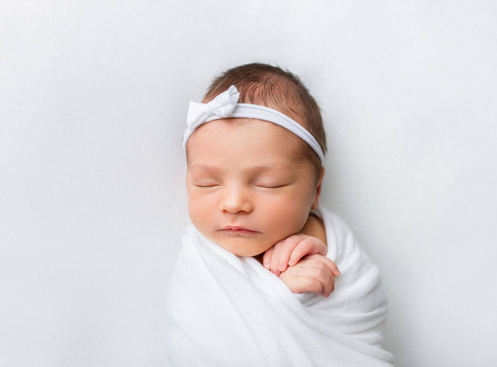 Newborn baby wrapped in white blanket, wearing white bow headband, eyes closed, resting.
