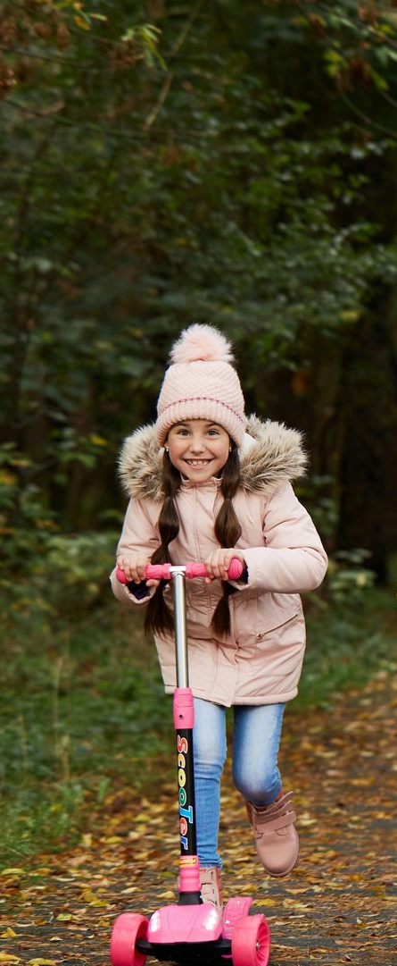 A smiling child riding a pink scooter on a path in a forest. She wears a pink coat and hat.
