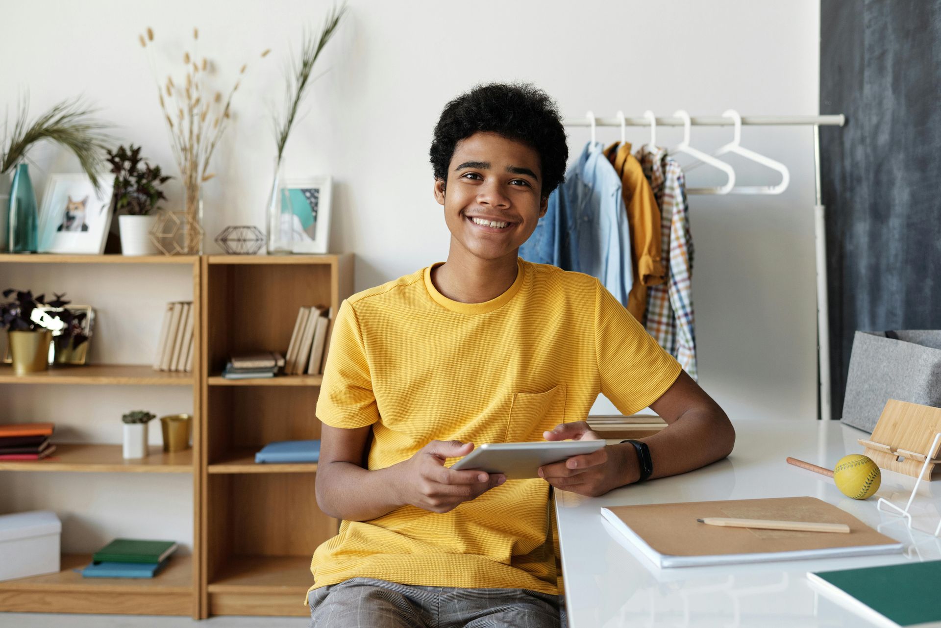 Teen smiles while holding a tablet at a desk. Yellow shirt. Shelves and clothing rack in background.