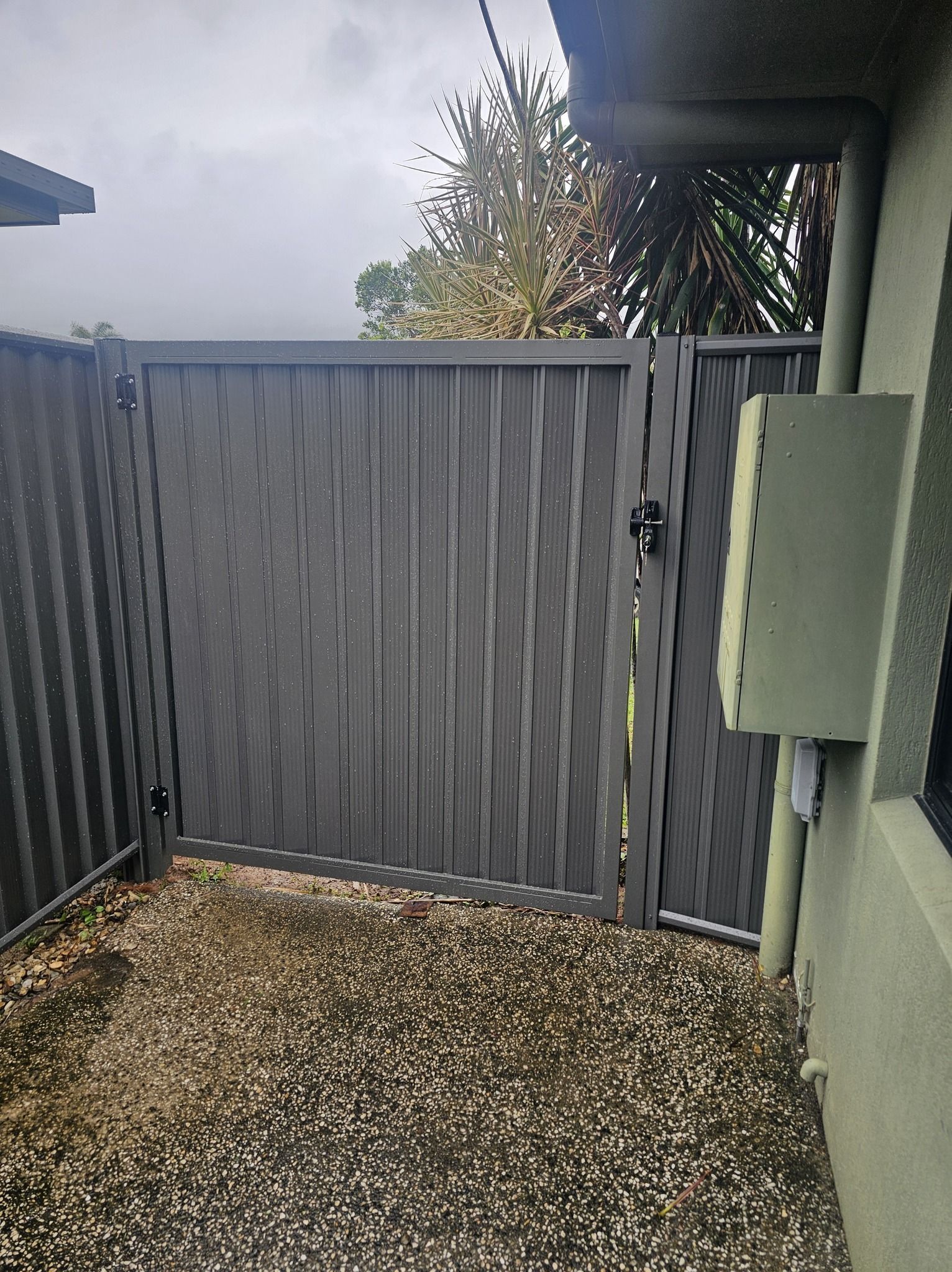 Narrow outdoor patio with a gray corrugated gate, gravel ground, and a wall-mounted utility box. — KLS Fencing In Edmonton, QLD