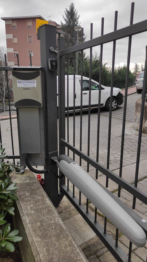 Cancello automatico con motore, centralina di controllo e pannello di controllo su una recinzione in ferro battuto. Sullo sfondo si vede un furgone bianco.