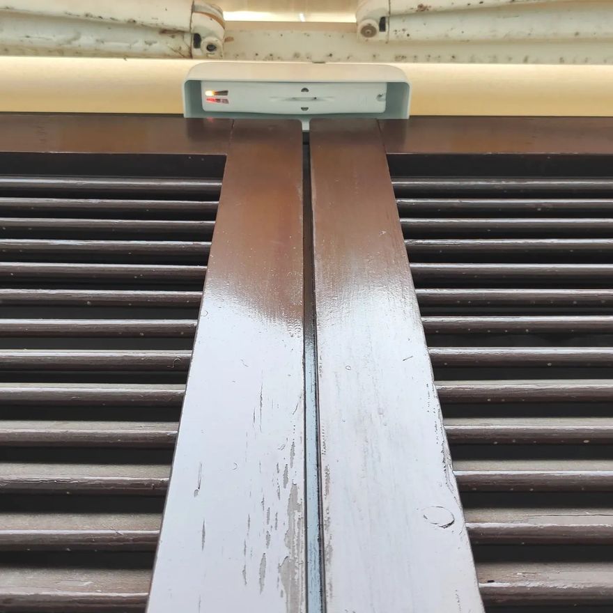 Persiane marroni con un dispositivo di sicurezza bianco  Wireless con Protocollo WLink nella parte superiore, una luce rossa accesa.