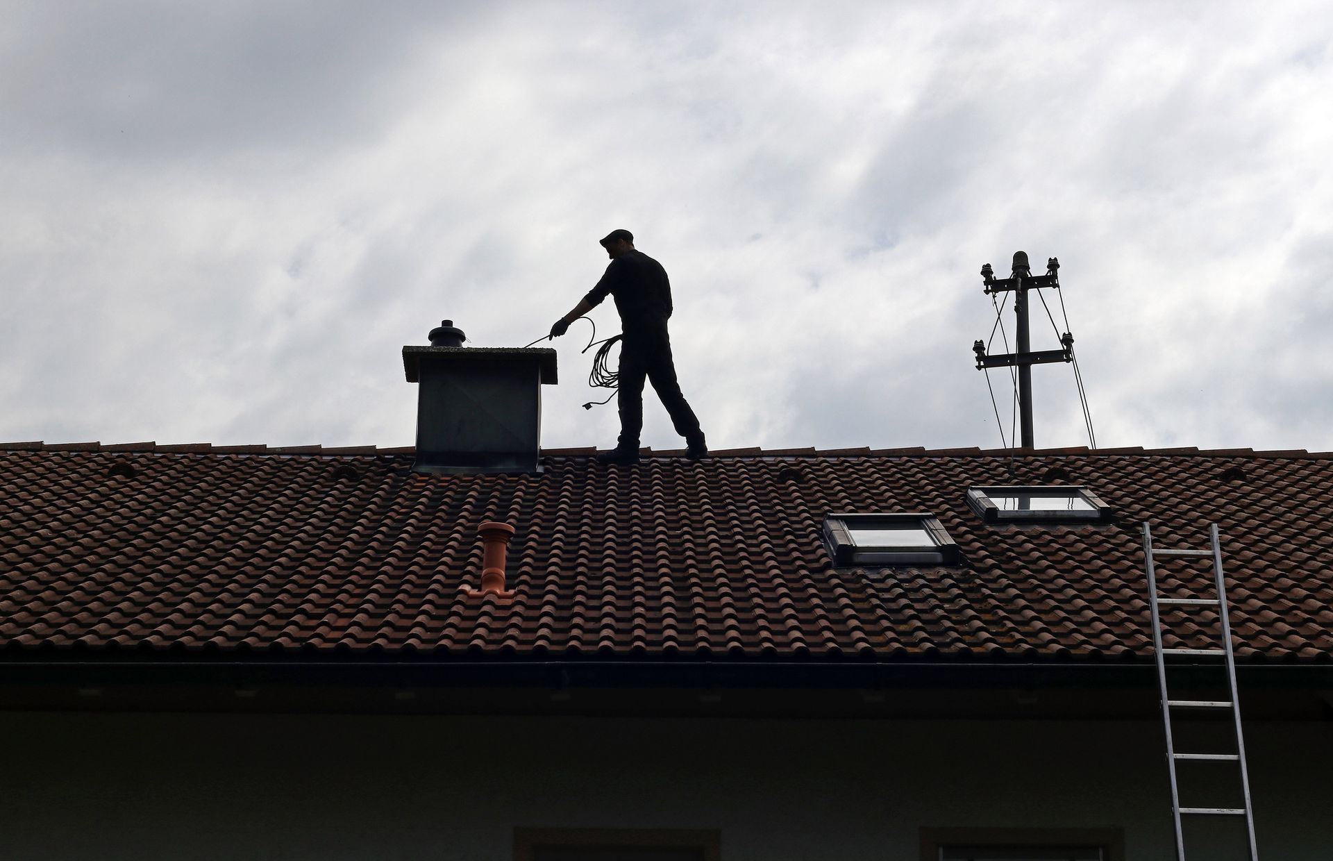 Silhouette of a person on a rooftop, possibly a chimney sweep, working near a chimney and ladder against a cloudy sky.