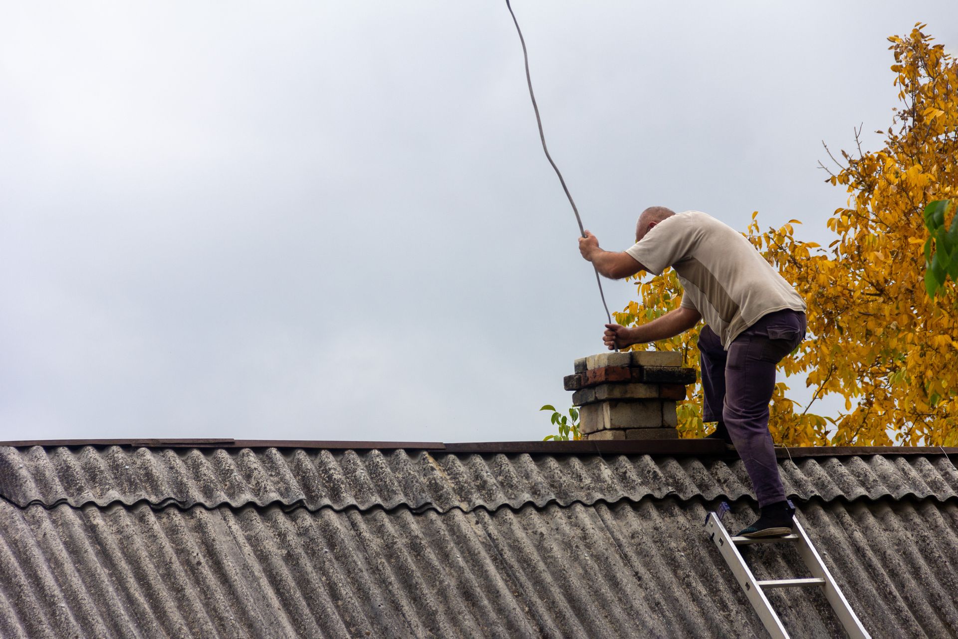 Man on ladder on roof, working on an antenna wire near a chimney, against a gray sky and fall foliage.