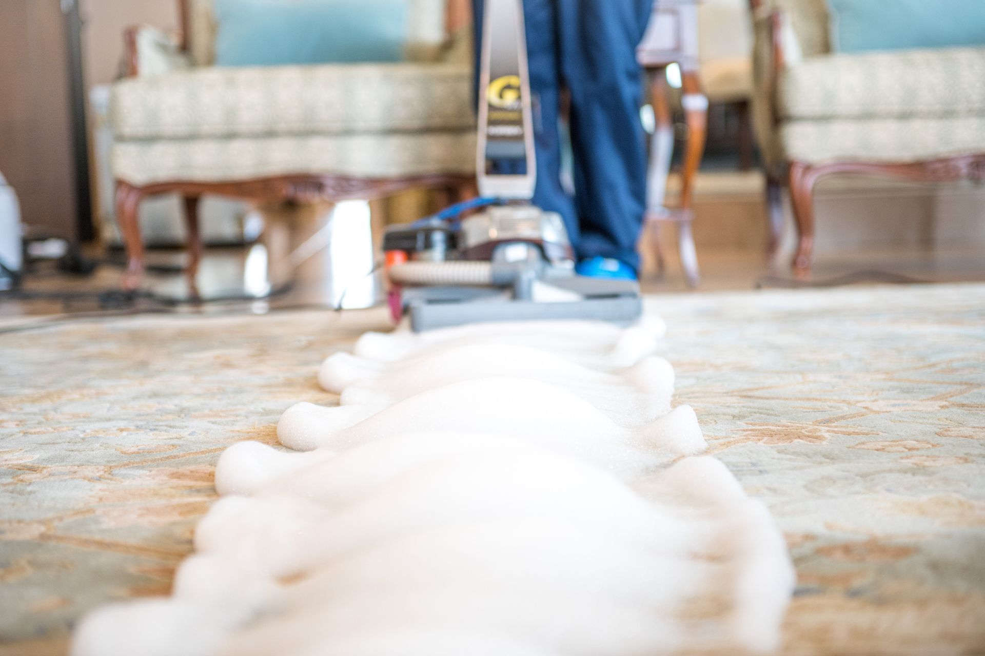 A person vacuuming a rug, leaving a foamy white cleaning trail. Beige rug, blue pants.