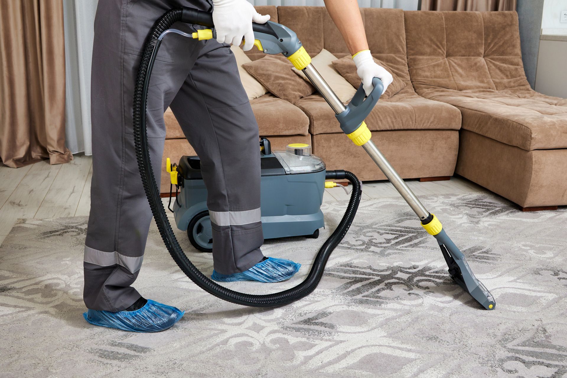 Person in gray uniform using a carpet cleaning machine on a patterned rug in a living room.