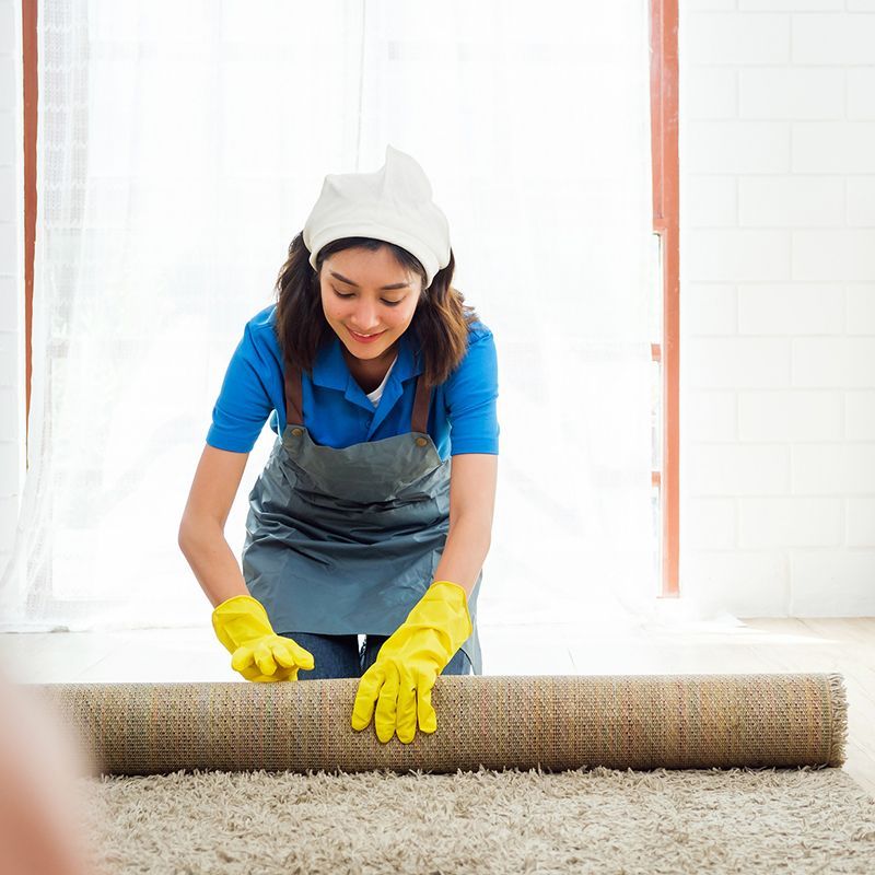 Woman in yellow gloves, rolling out a beige rug on a light-colored floor. She wears a blue shirt, apron, and head covering, smiling.