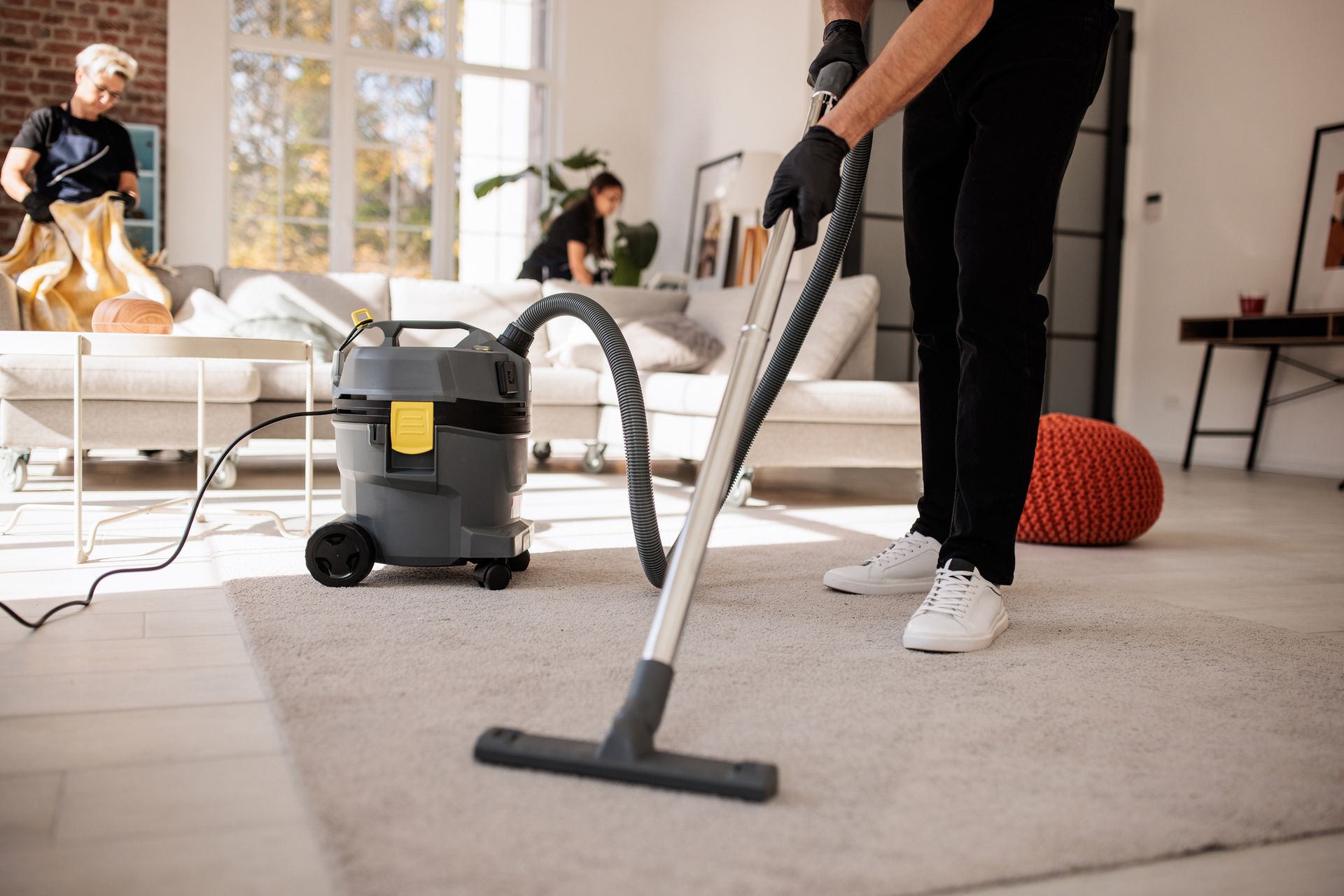 Person in black pants and gloves vacuuming a light-colored rug in a living room. Others are cleaning in the background.