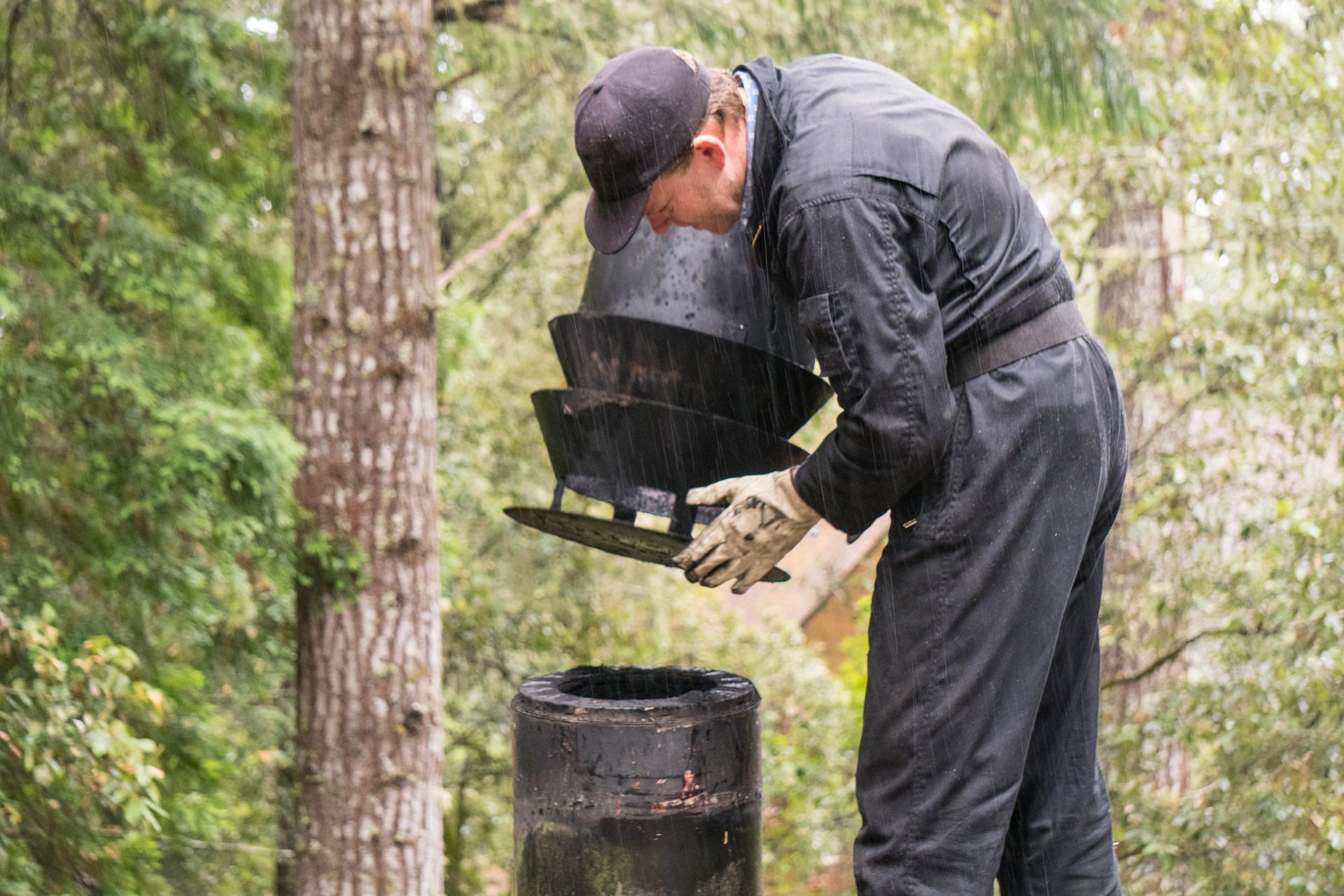 A chimney sweep, wearing black coveralls and cap, examines a chimney cap on a rooftop surrounded by trees.