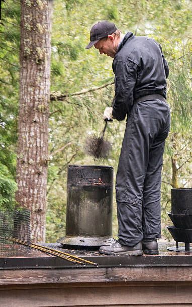 A chimney sweep on a rooftop cleans a chimney with a brush, wearing a black jumpsuit and cap.