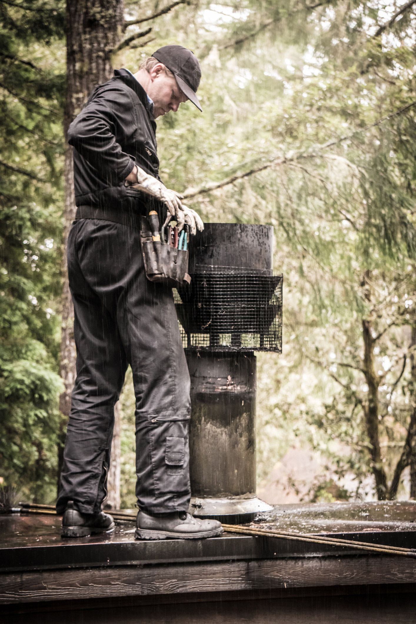 Man in work clothes cleaning a chimney on a rooftop, holding tools. He wears a cap, and the setting is outdoors among trees. Man in work clothes cleaning a chimney on a rooftop, holding tools. He wears a cap, and the setting is outdoors among trees.
