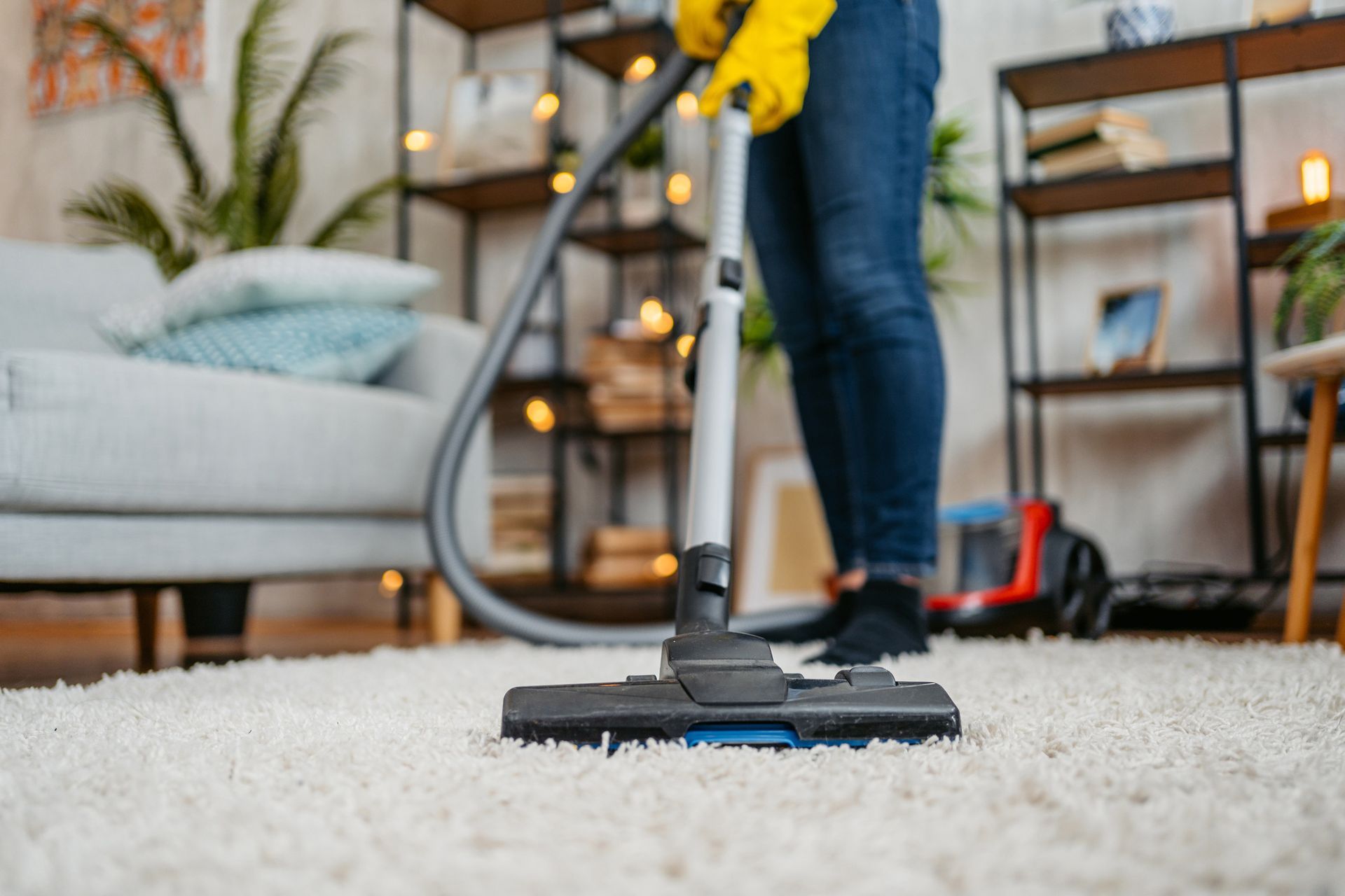 Person in jeans and yellow gloves vacuums a white carpet in a living room. Shelves and couch visible in background.