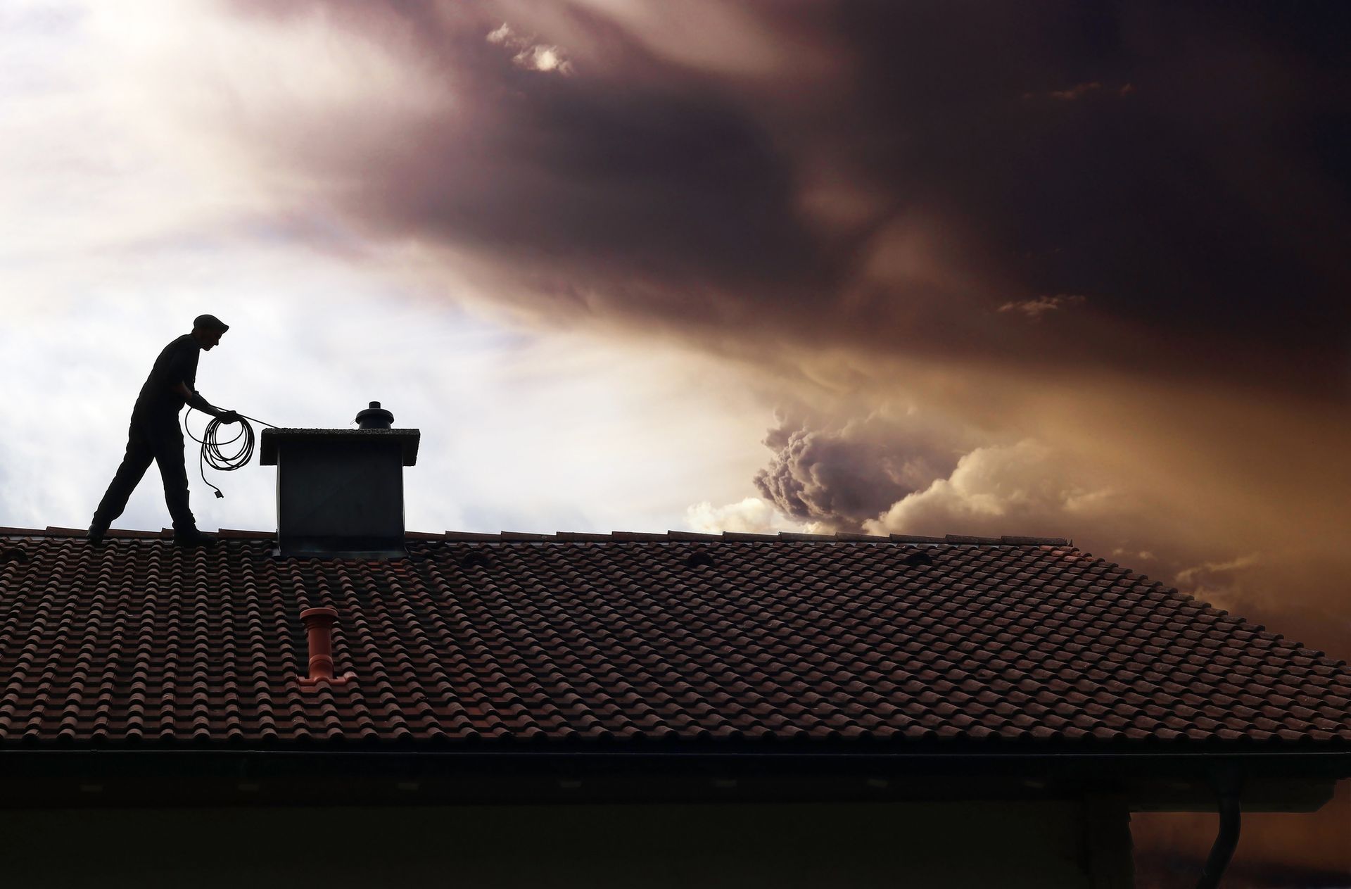 Silhouette of a person on a rooftop cleaning a chimney against a dramatic, stormy sky.