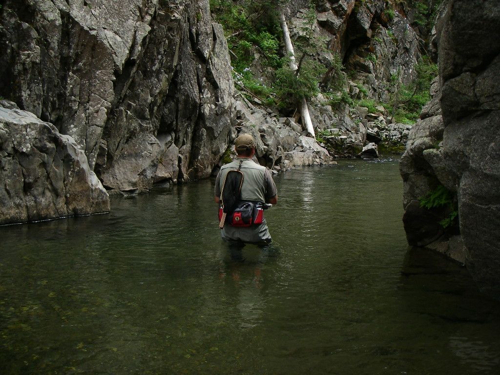 A man is standing in a river with a waterfall in the background.