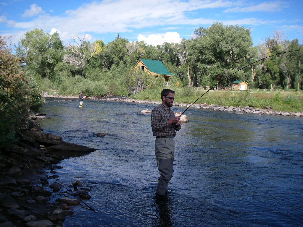 A man is fishing in a river with a house in the background.