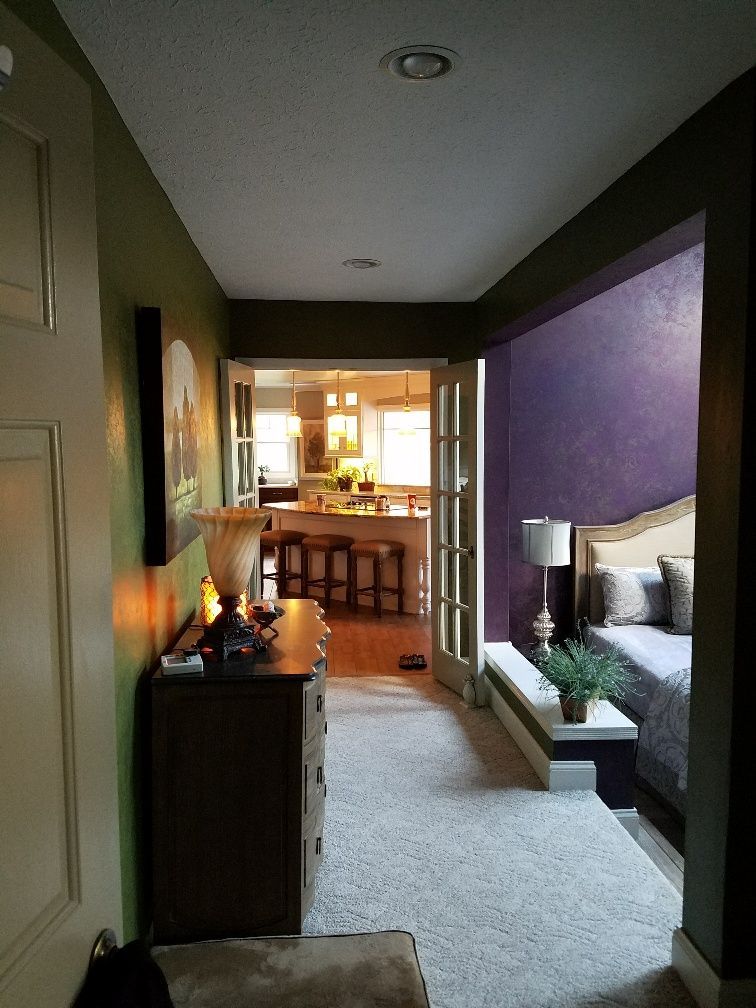 Hallway with chest of drawers, bed, and doorway to a kitchen. Olive, purple, and beige walls.